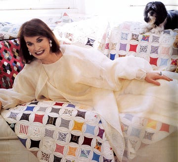 a smiling person lying on a bed with a little dog looking over from the top of the sofa