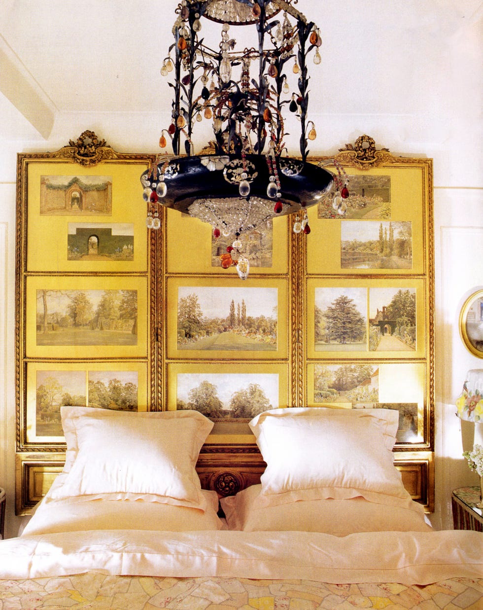 a bedroom with a chandelier of twisted iron and crystal and on the wall bedhind the bed is a screen with nine gold famed watercolors of landscapes