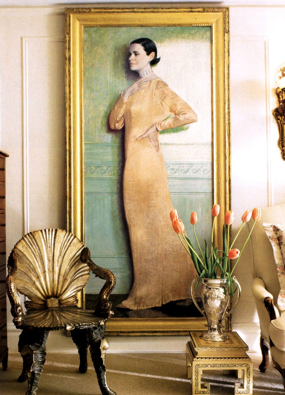 floor to ceiling gold framed painting of gloria vanderbilt in a long gold gown and slicked back hair and there is a shell back chair and some vases and low stools in front of the portrait