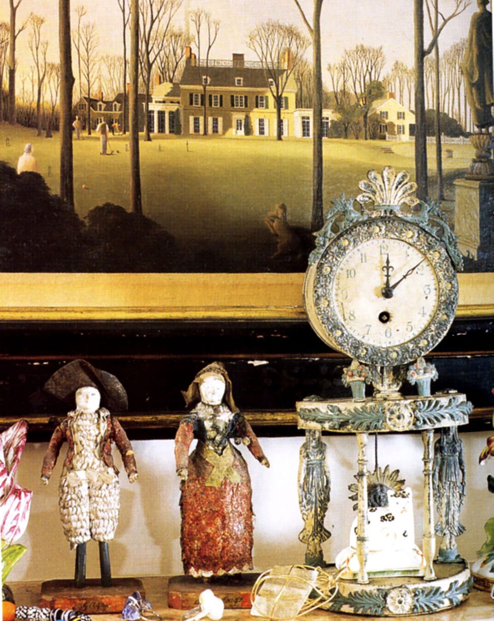 a clock and dolls on a table and a painting of a house above it on the wall