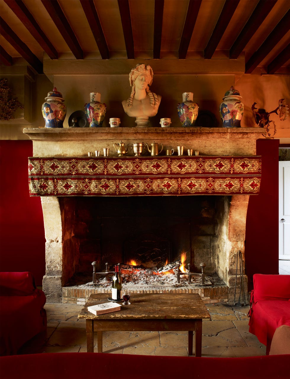 cozy living room with a large heavy stone fireplace and busts on the mantel and a red design runner on the front and a small cocktail table in front