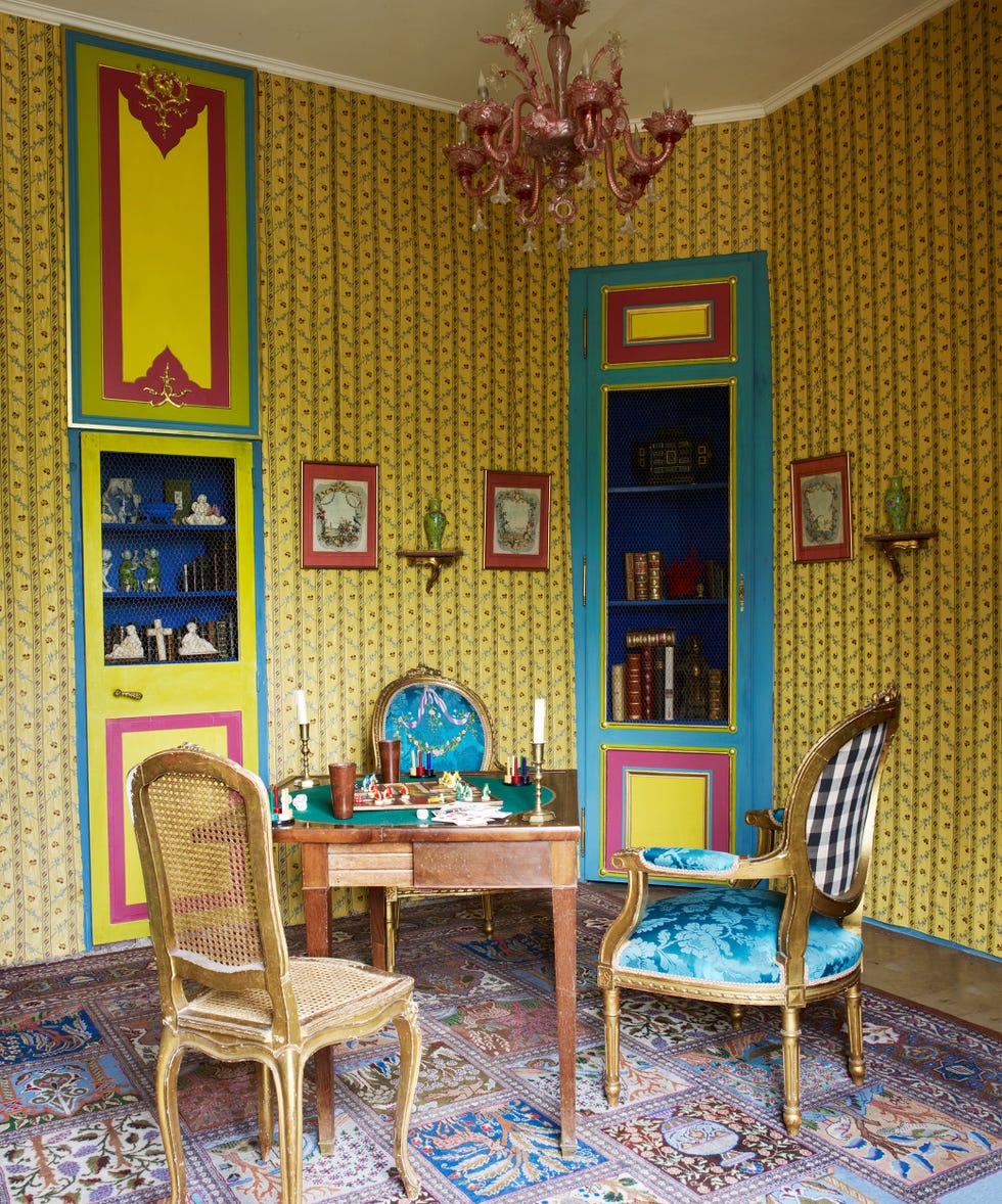 a colorful corner of a room with decorative yellow patterned wallpaper and inset shelves and hutch painted blue with small yellow doors and a gilded chair and a rattan back white washed chair and a block patterened rug