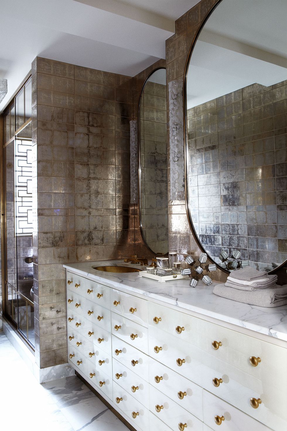 Bathroom with mercury glass effect tiles, large white vanity with brass pull handles and two large oval mirrors