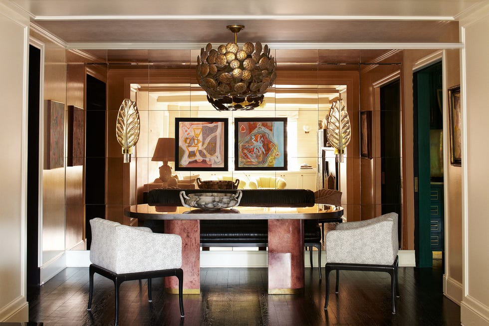 glamorous seating area with parchment and brass dining table top oval table and white boxy chairs and gold leaf shaped scones and a hammered gold light fixture over it