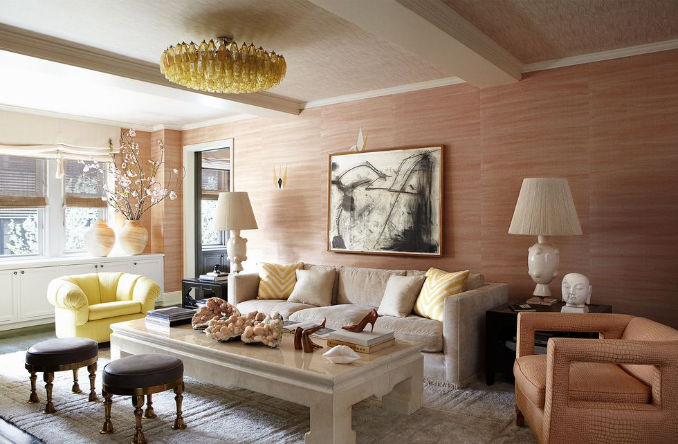 pink wallcovered spacious living room with a linen colored sofa and one open cut pink armchair and one yellow tufted side chair and blocky light colored marble like cocktail table and two small leather topped ornamental stools