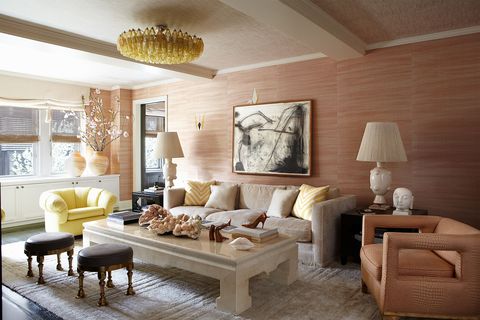 a spacious living room with pink wallpaper, a linen sofa and one pink open-cut armchair, one yellow tufted chair and blocky light marble like a cocktail table, and two small decorative stools with leather tops