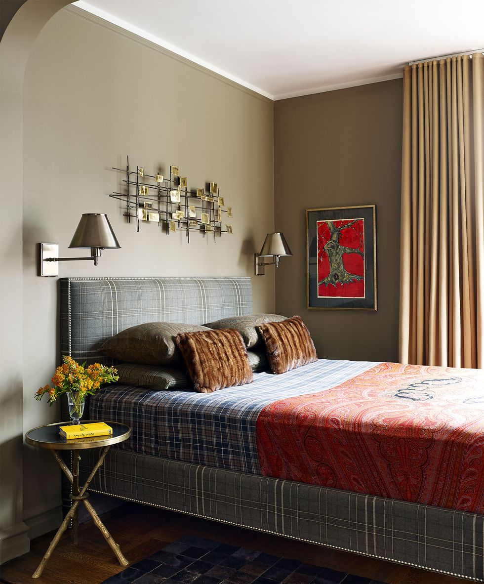 a bed with a gray suit type fabric on the base and headboard and a red blanket and an x leg gold rimmed side table topped with glass and a gold and silver abstract art above the bed and a window with long curtains