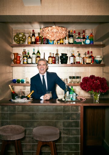 andy cohen stands in the bar area of his west village apartment, smiling wide and holding a frink