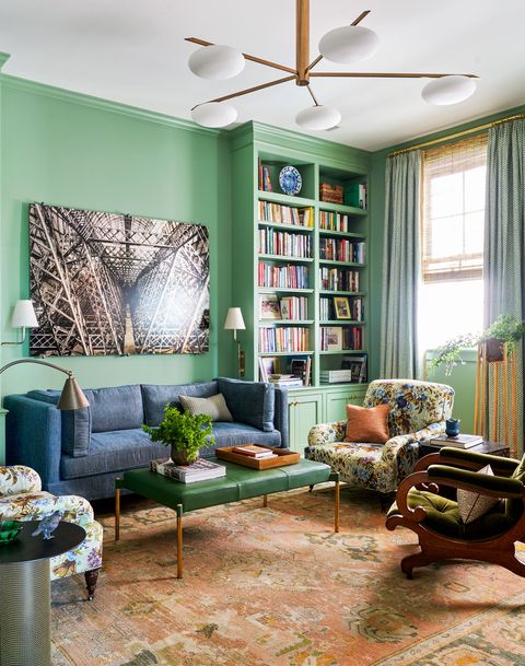 cozy library with mint green walls and built in bookshelves in the corner next to a window with complementary green curtains and  against the wall is a blue sofa with a large piece of art over it and a green leather bench used as a cocktail table and several chairs next to it on a patterned rug