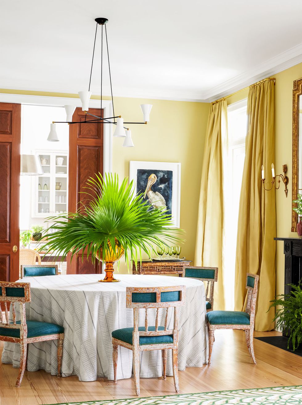 dining room in a soft yellow paint with darker matching floor to ceiling curtains on tall window and a round dining table at center with draped white tablecloth and green upholstered washed wood swedish chairs pulled up to it