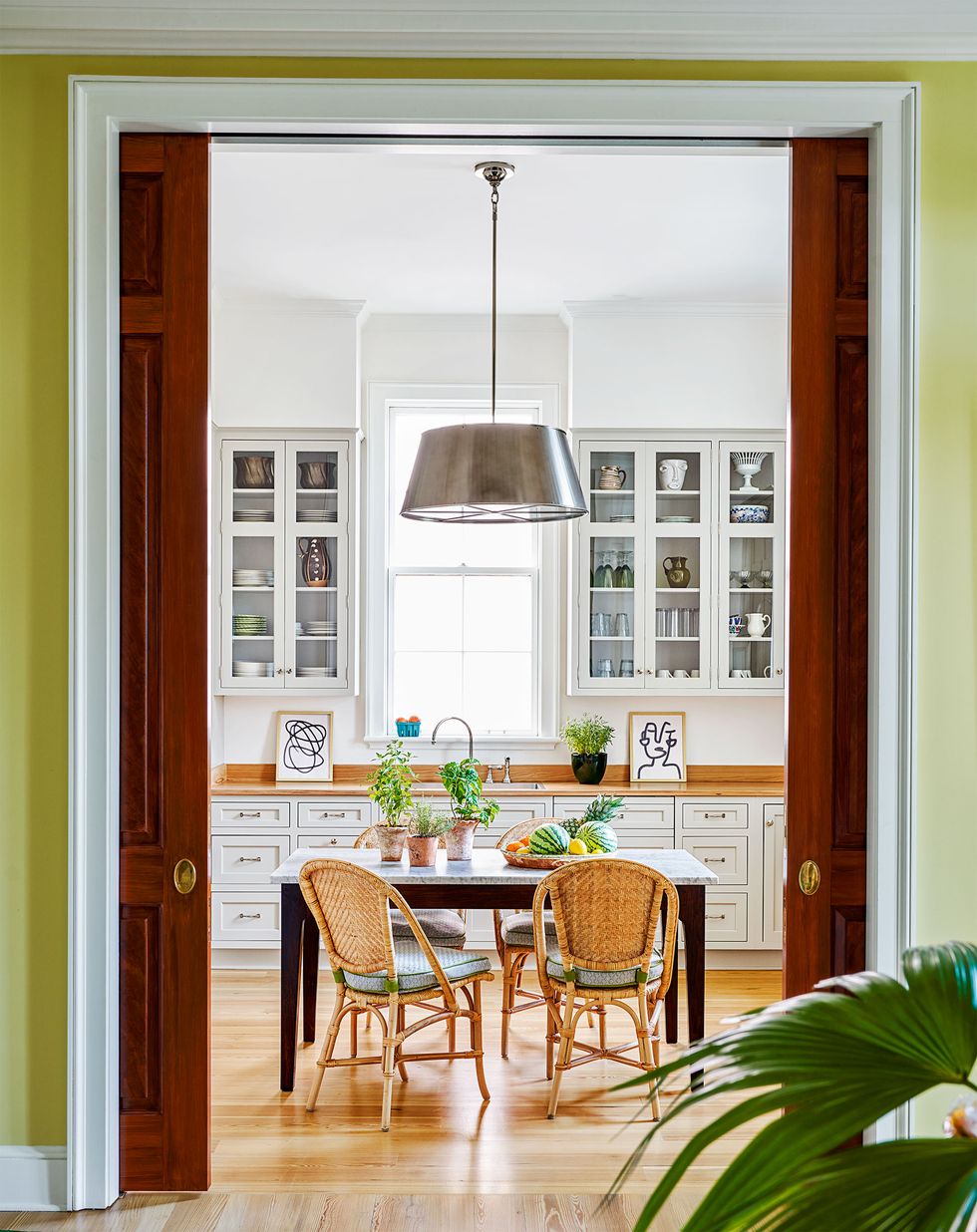view through a pocket doorway with yellow walls and white trim into the kitchen with a white wall cabinets and a small square table with rattan chairs pulled up to it