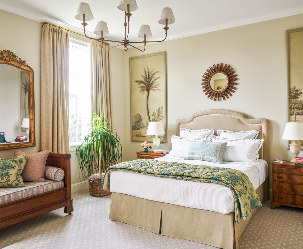 a primary bedroom has off white walls, a beige  upholstered bed, mahogany night stands and daybed with cushions and a gilt mirror above it, lightly patterned carpet, chandelier with five shades, curtained window
