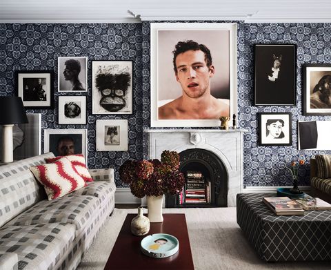 living room with mantle and large photograph of a young man over the fireplace against a blue and white patterned wallpaper and many other framed images and a sofa with large box pattern in gray and light tones and a cocktail table with flowers