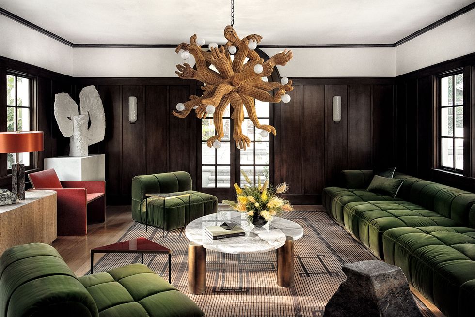 dark wood panelled living room with chandelier made of multiple arms and hands holding lights and a large green velvet tufted armless sofa and chairs and a low cocktail table at center with marble top and wooden legs