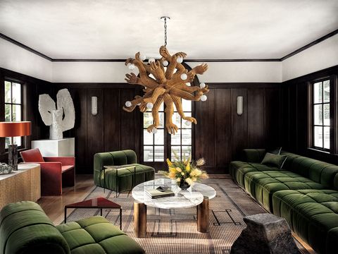 dark wood panelled living room with chandelier made of multiple arms and hands holding lights and a large green velvet tufted armless sofa and chairs and a low cocktail table at center with marble top and wooden legs