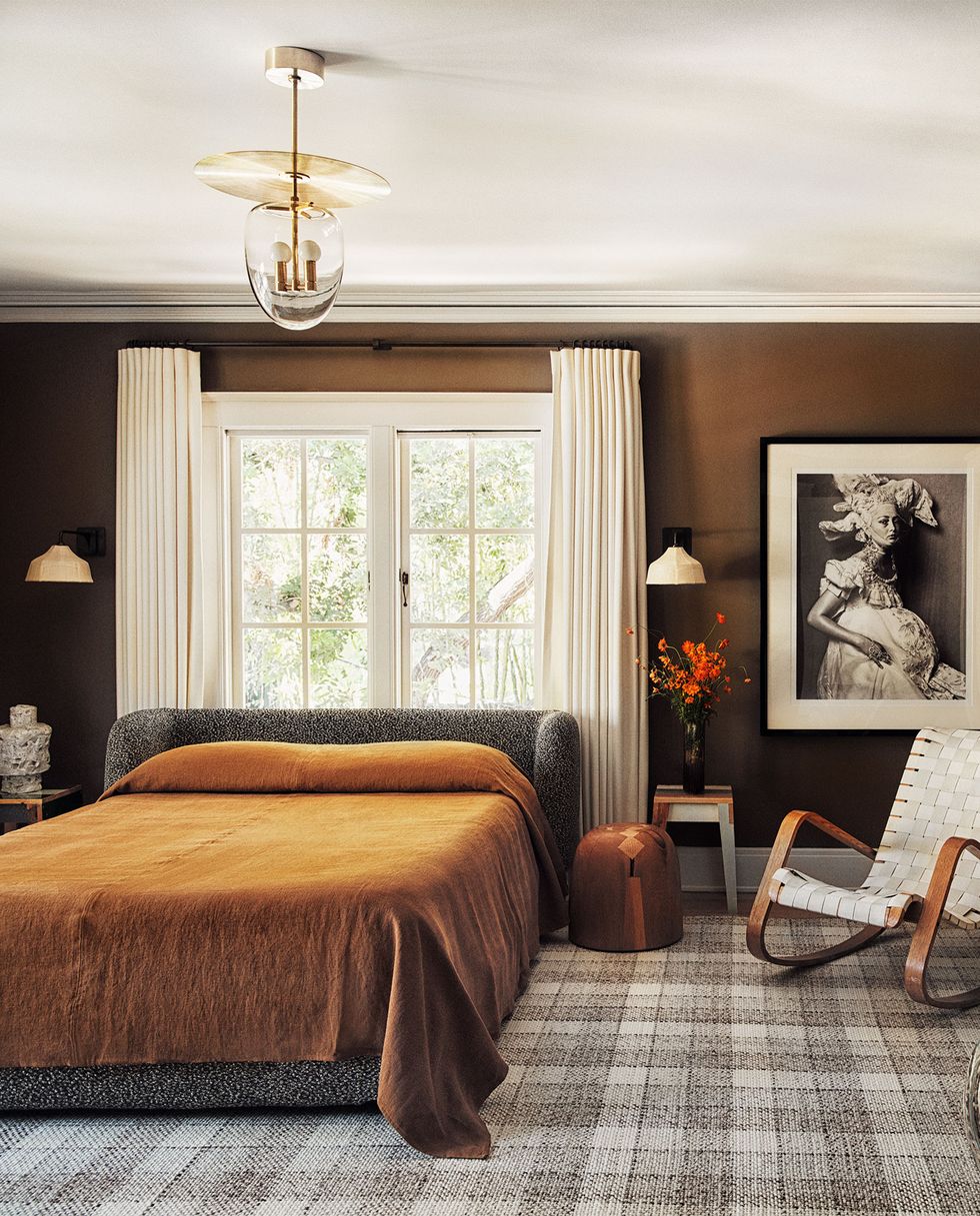 primary bedroom with medium chocolate painted walls and a plaid rug in neutral tones and a slatted rocking chair next to the bed with complementary coverlet in brown on the bed