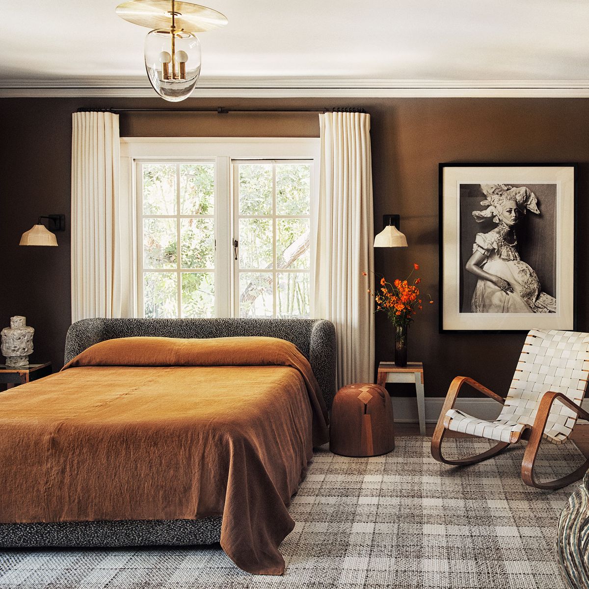 primary bedroom with medium chocolate painted walls and a plaid rug in neutral tones and a slatted rocking chair next to the bed with complementary coverlet in brown on the bed