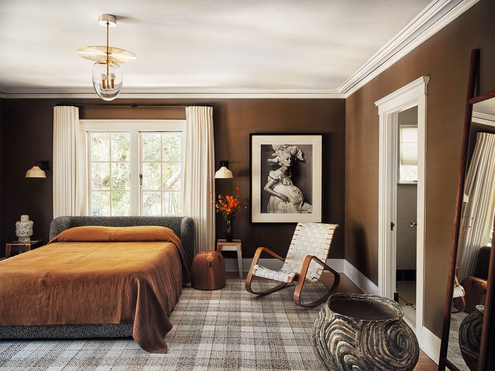 chambre principale avec des murs peints en chocolat moyen et un tapis à carreaux dans des tons neutres et une chaise à bascule à lattes à côté du lit avec une couverture complémentaire en marron sur le lit