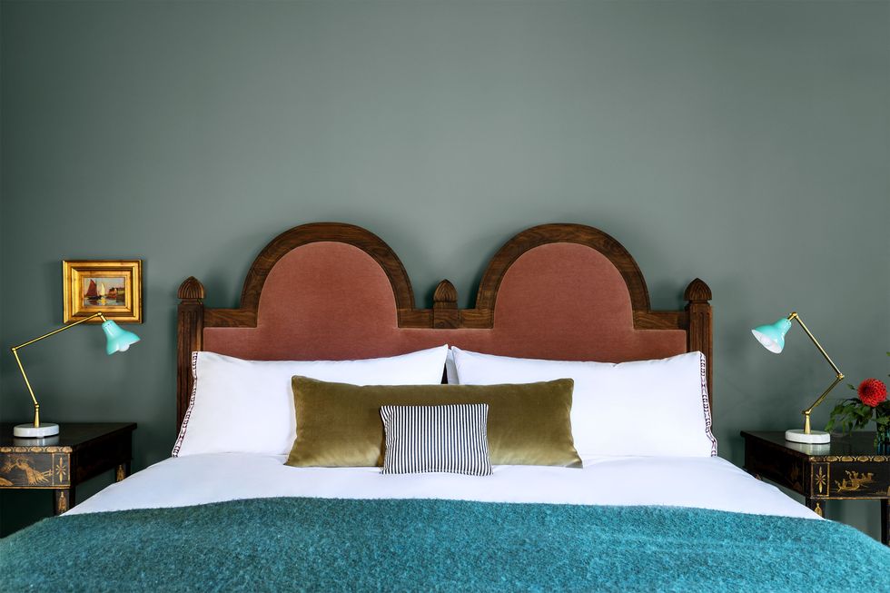 bedroom with muted green painted wall and bed with double hump wooden headboard upholstered at center in a dark salmon velvet and a bright turquoisy nubby coverlet