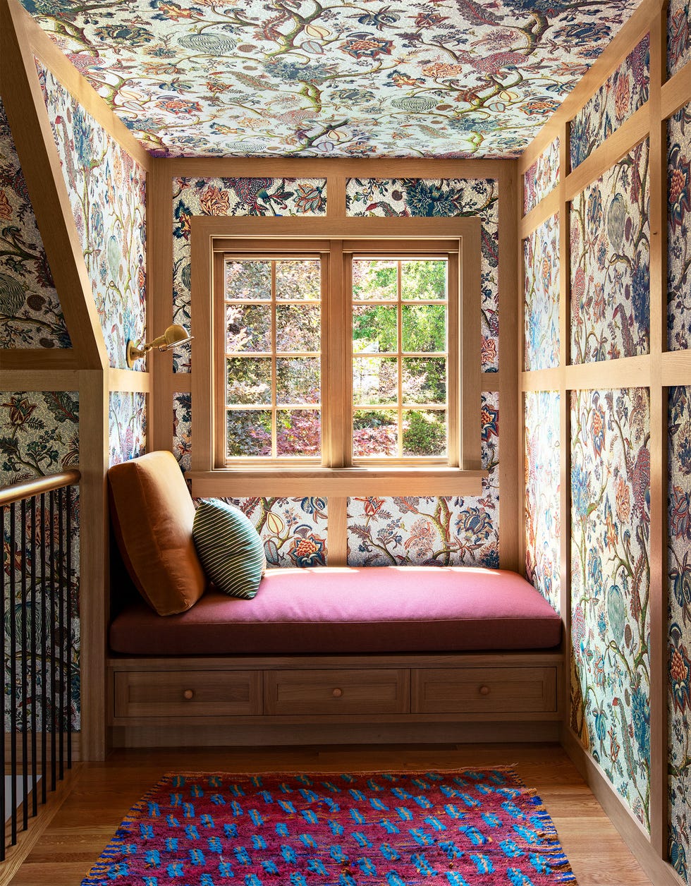 daybed in a nook in a wood panel frame window with bold flower print wallpaper plum colored rug with blue specs and a salmon cover on the bench