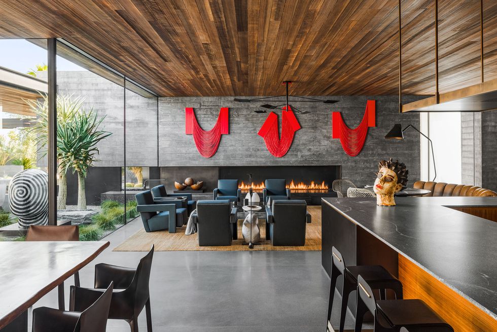 open kitchen dining living area great roomwith long black counter and stools and a gray brick wall with modern fireplace with fire in it and three large red pieces of artwork above it and a wood paneled ceiling and floor to ceiling glass walls on the left