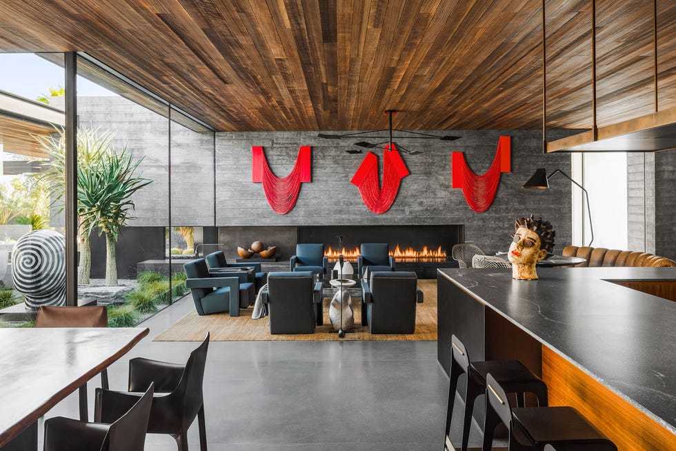 Studio AR&D - ELLE DECOR open kitchen dining living area great roomwith long black counter and stools and a gray brick wall with modern fireplace with fire in it and three large red pieces of artwork above it and a wood paneled ceiling and floor to ceiling glass walls on the left
