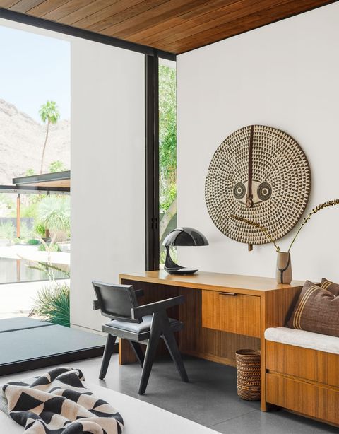 office slash guest room with a teak desk and attached daybed with a black chair pulled up to the desk and an african mask on the wall above and wall of glass to the left