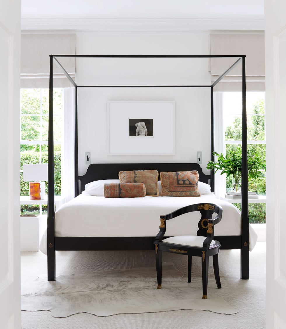October 2023 - ELLE DECOR between two windows sits a black four poster bed with white bedding and colorful pillows, white walls, a framed black and white photo, white carpet and a white cow hide rug, and an antique empire chair