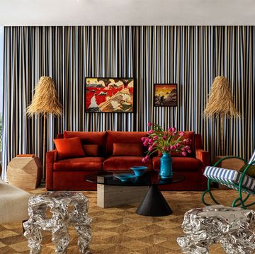 a living room has a striped curtain along the back wall, a burnt orange sofa, two floor lamps with thrush shades, a rocking chair with green metal base and striped cushions, two amorphous silver side tables
