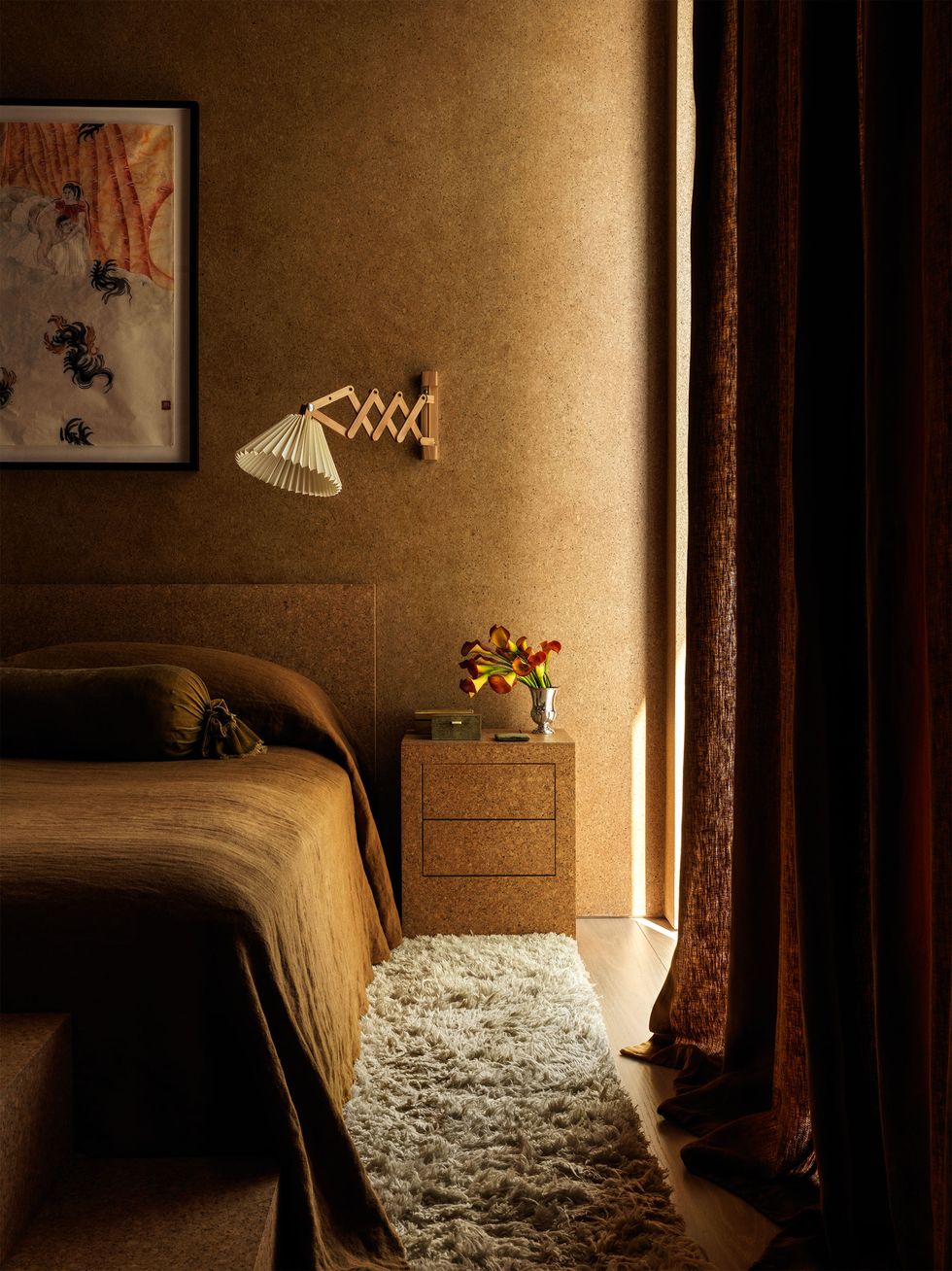a bedroom has cork wallcovering and a cork headboard and nightstand, a sconce with an accordion arm and pleated shade, brown bedcover and curtain, a light shag rug, and a painting over bed