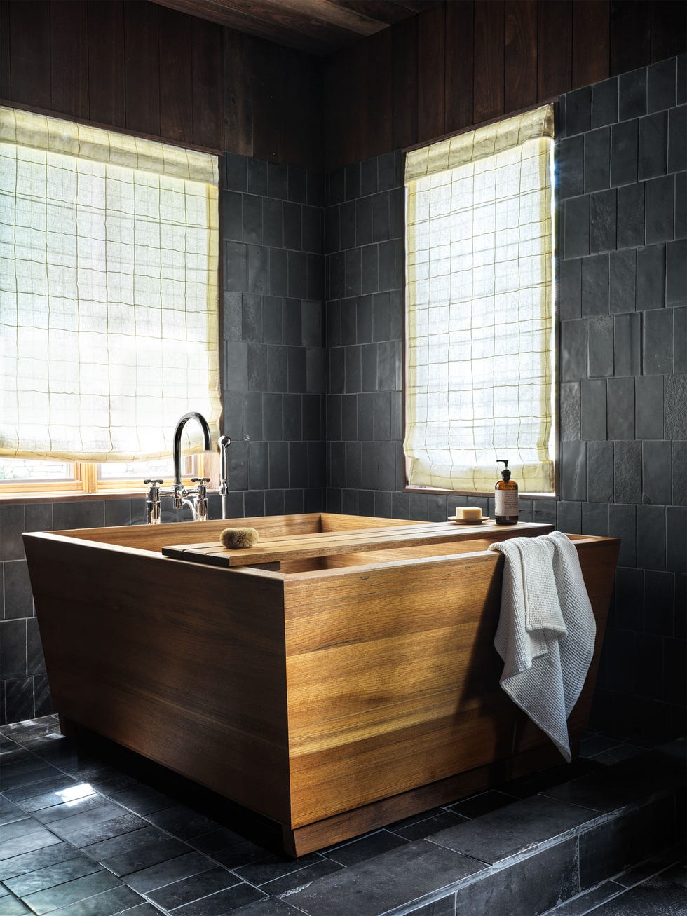 a bathroom has black limestone tiles on the floor and walls, two windows with linen roman shades in a small square pattern, a deep square cedar tub with a wooden tray holding soap and a dispenser, chrome fittings