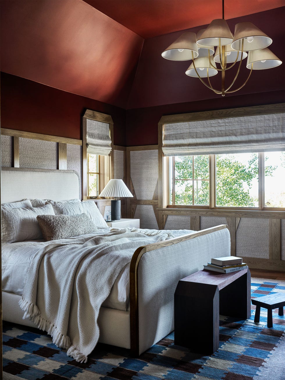 Electric Bowery - ELLE DECOR a bedroom has deep red upper walls and ceiling, linen covered wall panels separated by wood slats, windows with linen roman shades, a bed with fabric head and footboards, a blue, brown, and beige checkerboard rug