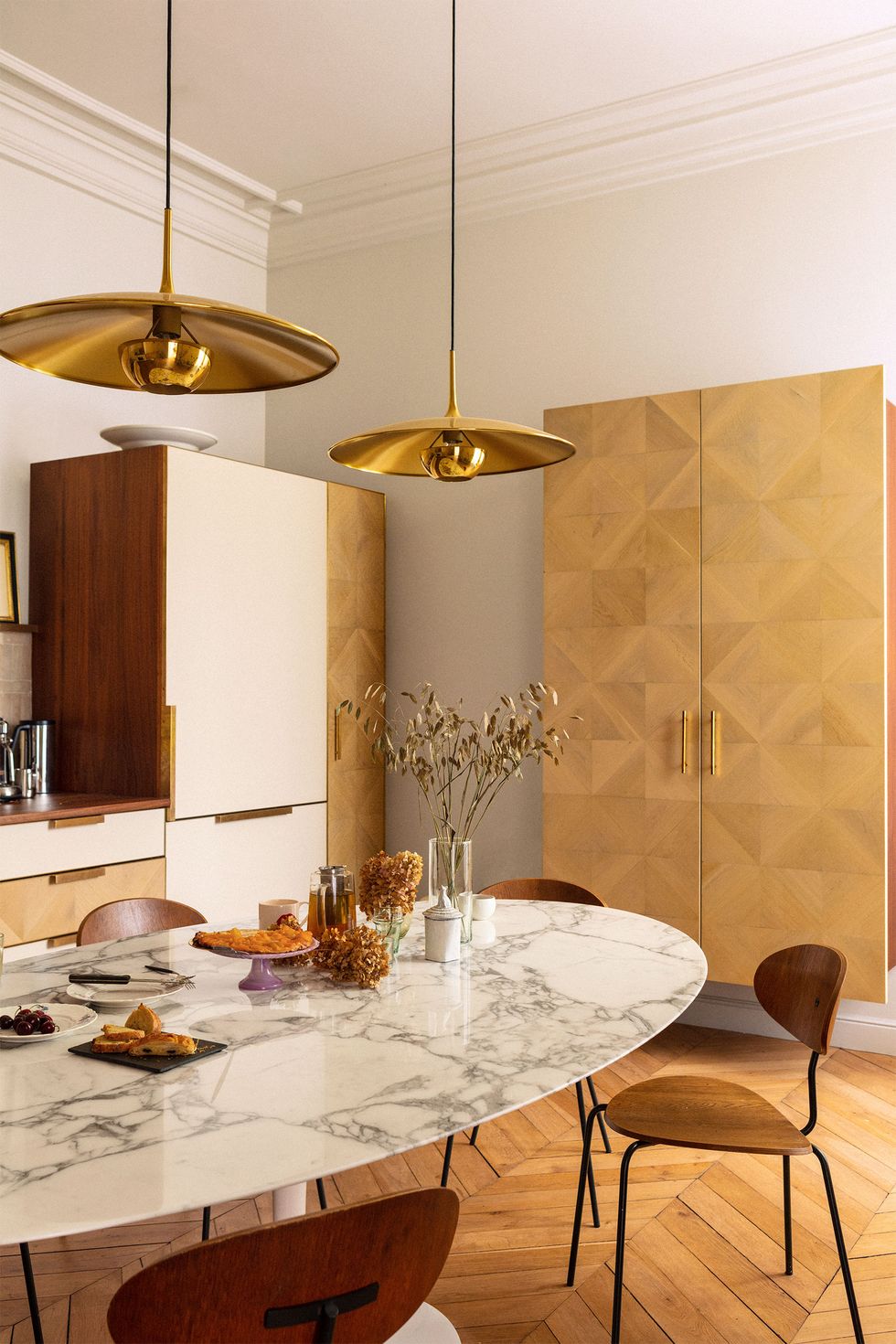 eat in kitchen with chevron design wood floor and an oval white marble looking thin pedestal table with klimos like chairs and gold pendant hood lights and a long golden colored suspended cabinet at the back wall