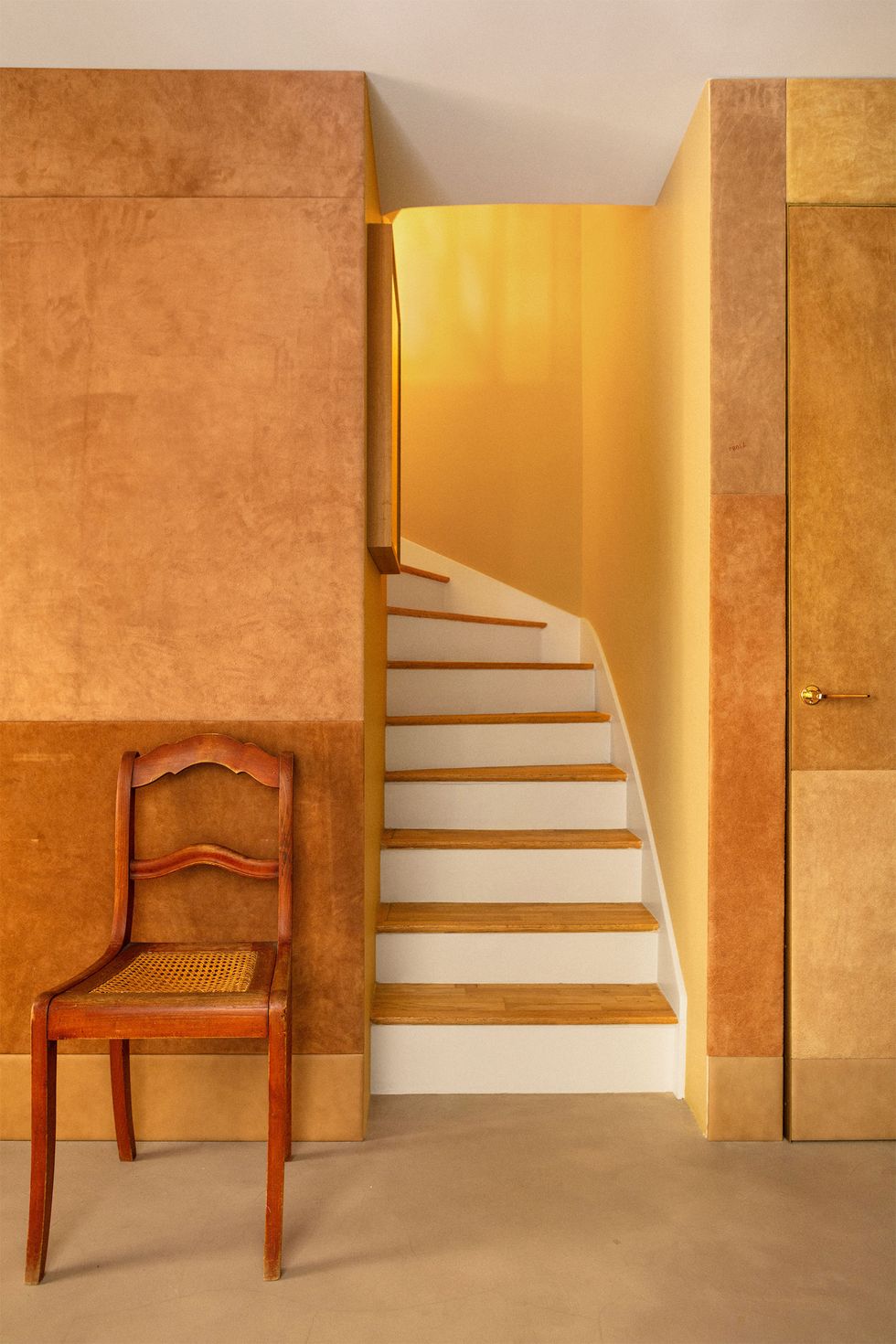 light colored space with honey colored walls and floor a small stairway leading up and a beautiful old cane seat chair standing guard