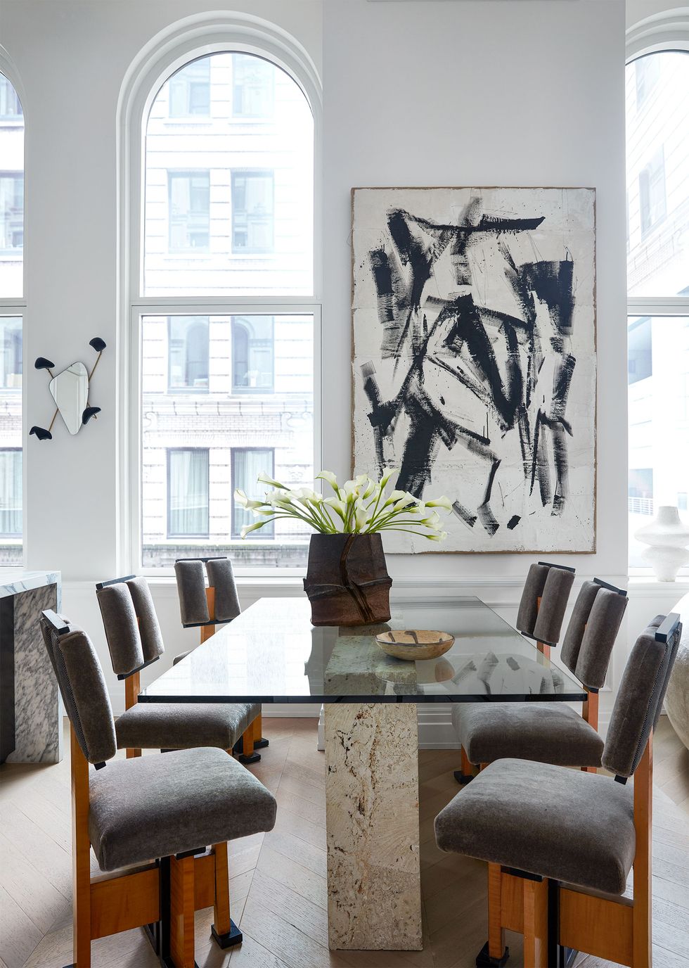 a room with a marble pedestal base table and gray suede chairs with wood frames and a black and white abstract painting on the far wall next to window