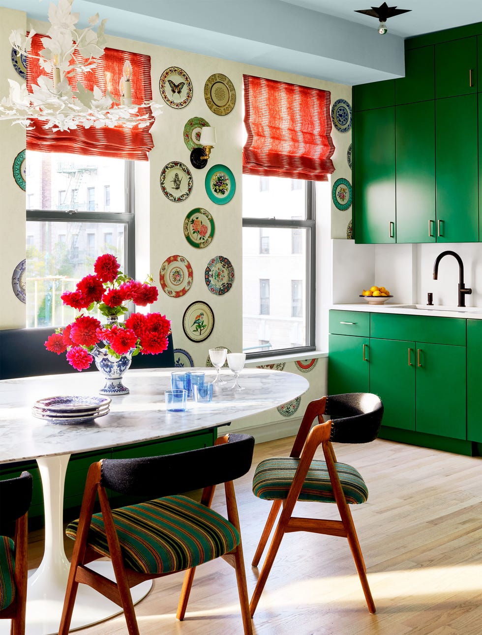 kitchen with bright green cabinets, white tile backsplash and dark faucet, wallpaper with white background and colored decorative plates, marble oval tulip table, wood chairs with green striped seats, red window shades