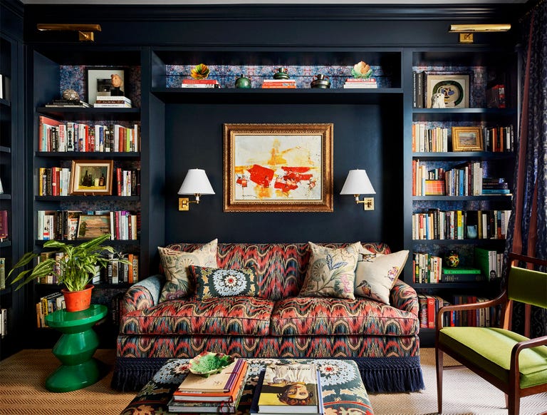 library with dark blue walls, built in bookshelves on both sides of a sofa with a red and black wave pattern with black bottom fringe, sconces and artwork above sofa, green side table, print fabric cocktail table, green chair