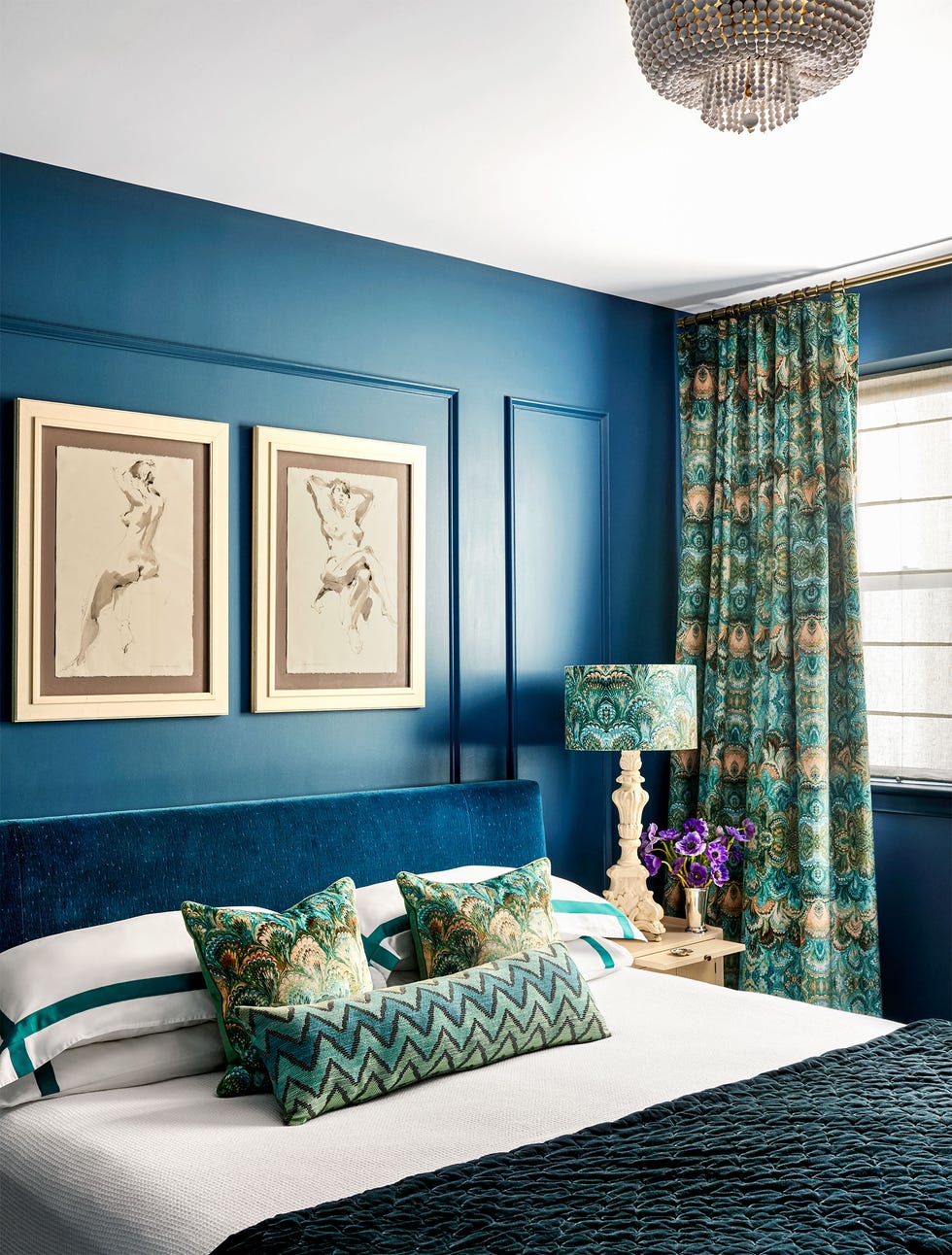 deep blue walls, bed with blue velvet headboard, white bedding, green hued patterned accent pillows and throw, nightstand with ornate lamp, curtains and lampshade match pillow fabric, framed drawings