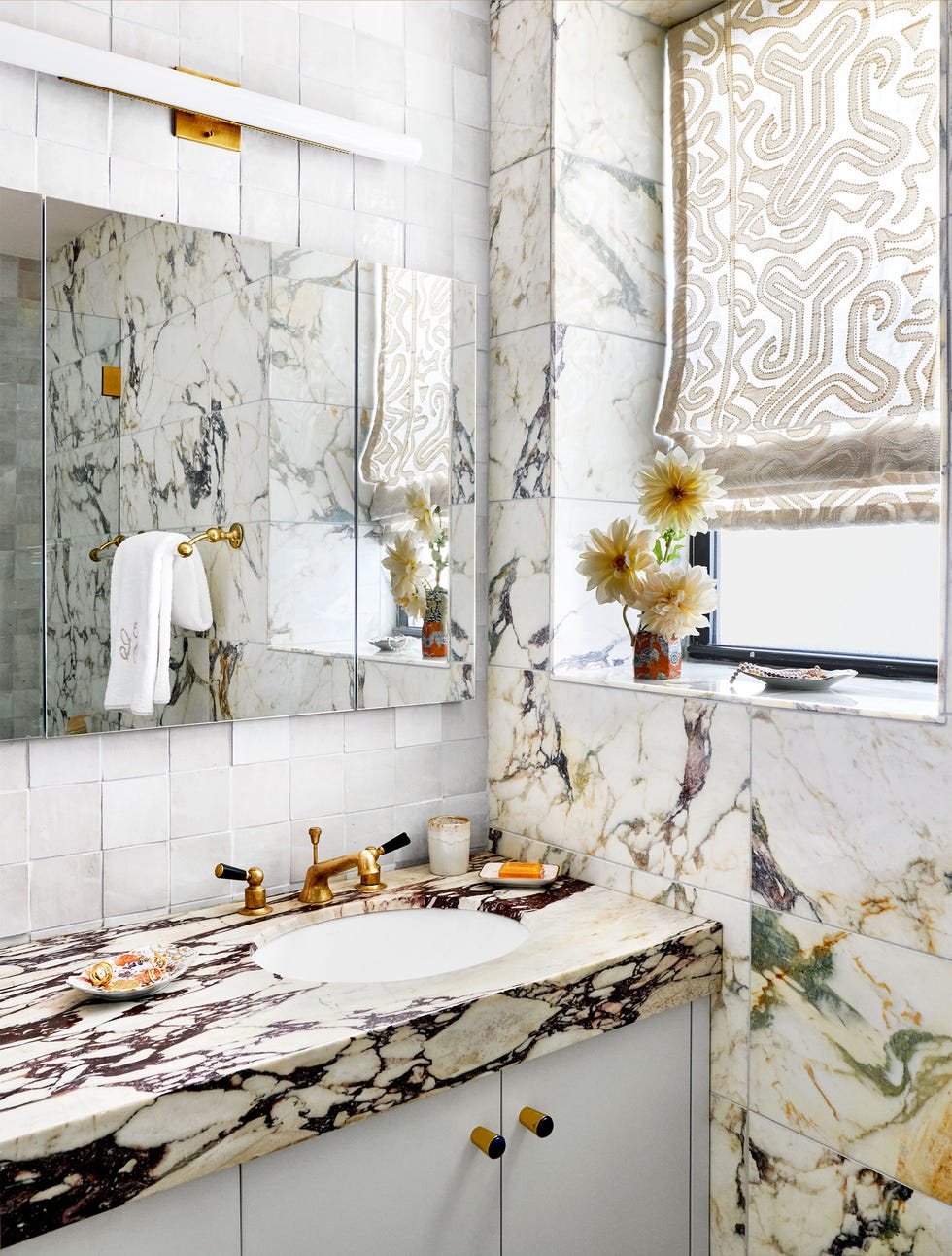 bathroom with violet veined marble sinktop and walls, white under cabinets, square white tiled backsplash, brass fittings, large mirror reflecting small brass towel holder with white towel, patterned fabric roman shade