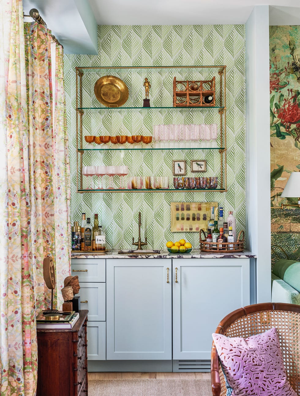 bar with green patterned wallpaper above light blue cabinets and drawers, marbled topped counter with sink, two trays of spirits, bowl of lemons, three glass shelves above with glasses and art objects, print curtains,