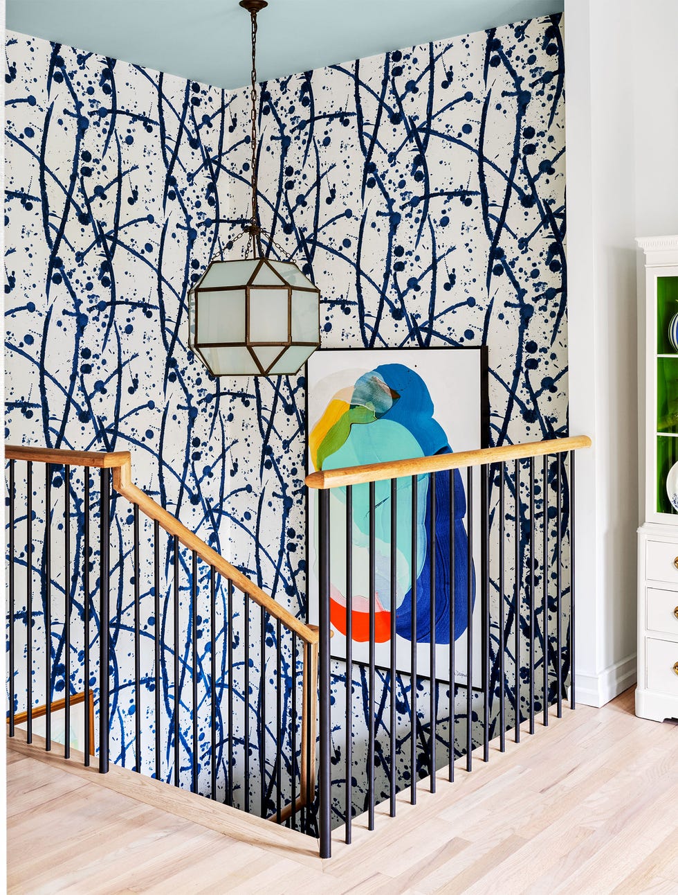 stair landing with light wood floor, wood banisters and plain black balustrade, walls covered in a white with deep blue splattered design wallpaper, colorful framed artwork, frosted glass pendant