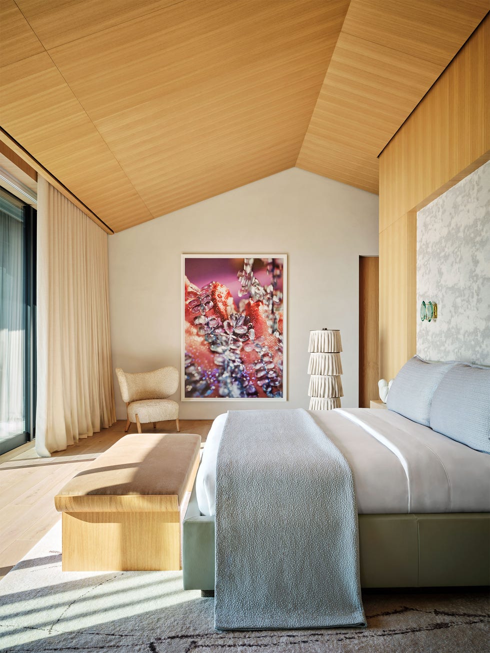 Rafael de Cardenas - ELLE DECOR a bedroom with light full length curtains, a white wall and a wood paneled ceiling and wall with wallpapered indent above the bed, chair in corner, floor lamp with pleated shades, colorful artwork, rug
