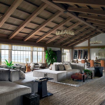 Steven Gambrel - Tom Kligerman - ELLE DECOR large great room with two clusters of gray sectionals, club chairs, cocktail tables, dining table and chairs, textured rug, two multilight pendants, pitched beamed ceiling, large window with seaviewsteven gambrel tom kligerman elle decor