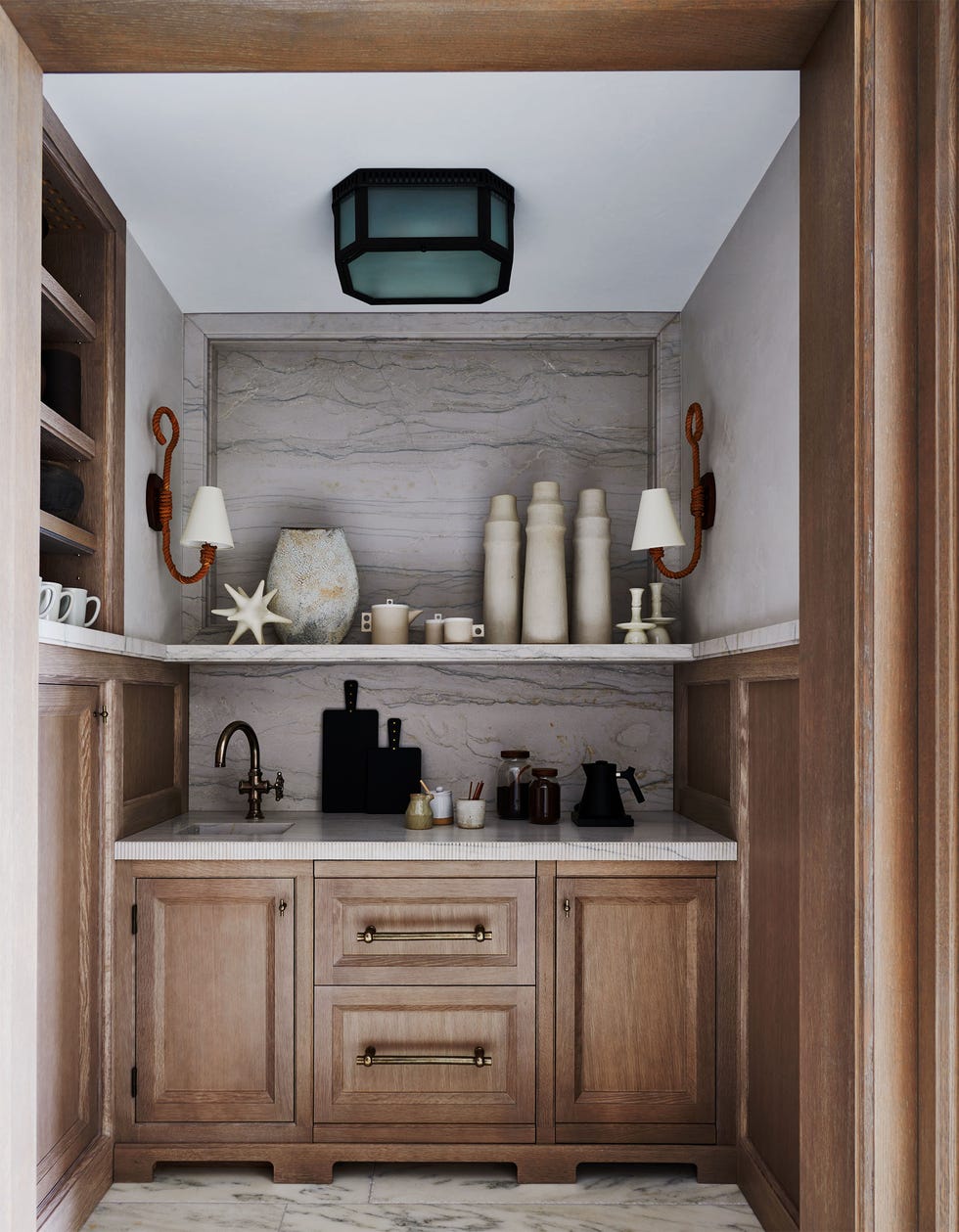 a nook with marble floor and back wall, small counter with a sink and coffee pot, wooden cabinets and drawers below, shelf above holds ceramic carafes, a vase, two sconces, built in wood shelves with cups