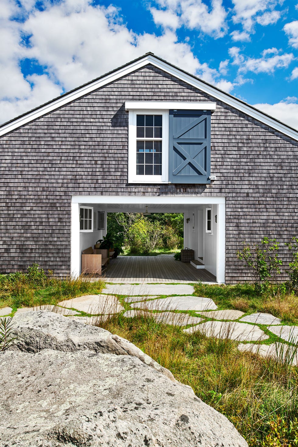 exterior of house has weathered cedar shingles, pitched roof, center of structure is open with wood floor, wood bench and an entry door, above is a large window with sliding door, rocks and grasses in front