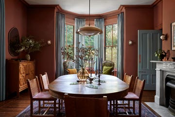 dining room with dark terra cotta walls and light blue gray curtains and paint trim, oblong wood table with chairs with leather seats, pendant above, fireplace with ornate surround, carved wood sideboard, patterned rug