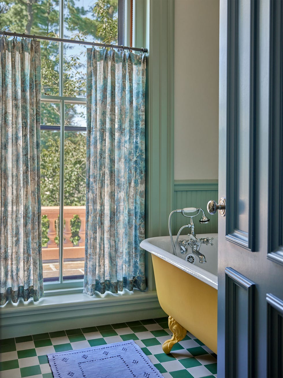 soft white bathroom with sage trimp and wainscoting, claw foot tub painted mustard yellow, silver fittings, large window with translucent floral curtains, green and white checked floor, blue runner, blue gray door