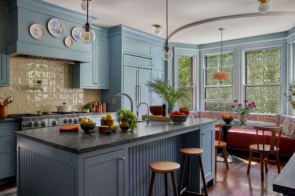 kitchen with baby blue walls and cabinets, white tiled backsplash, decorate plates, marble topped island with sink, two globe pendants, barstools, bay window with table and inset bench and chairs, rattan pendant