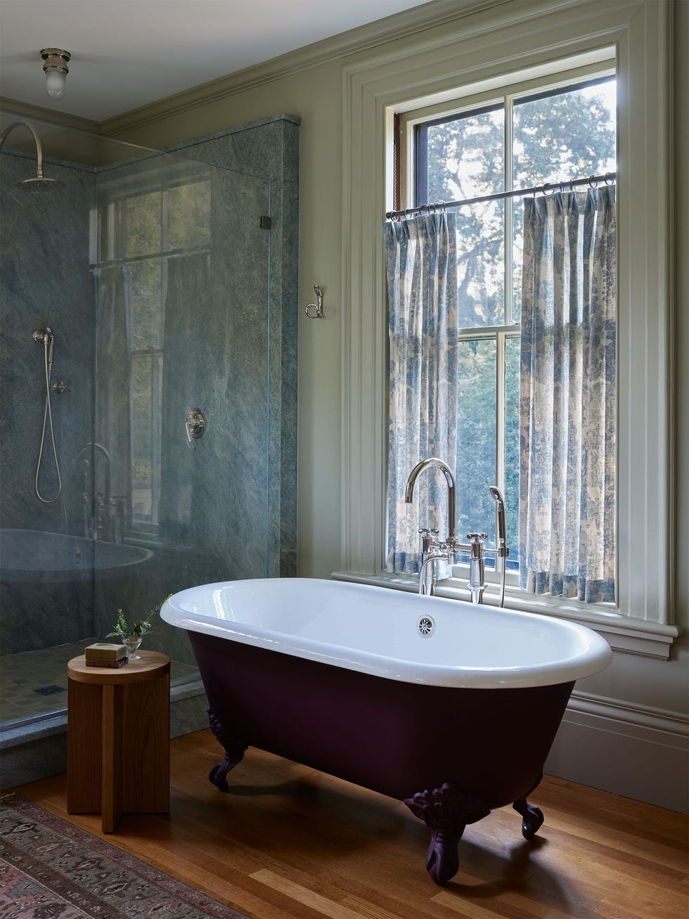 bathroom with a claw foot tub with a deep purple painted exterior, silver fittings, round wood table, window with translucent flowered curtains, shower with marble walls and glass door, wood floor