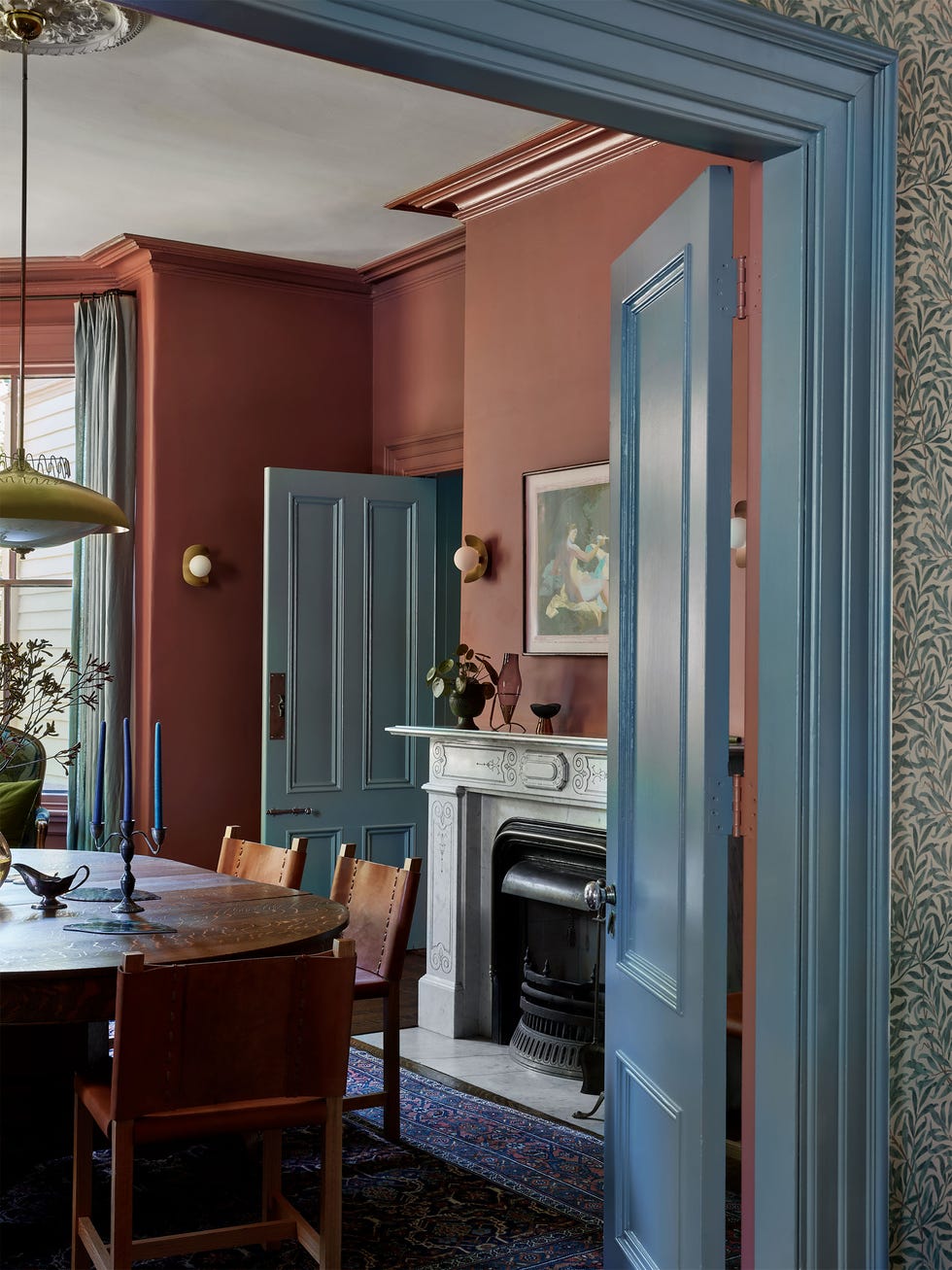 entry to dining room with deep terracotta painted walls and blue gray doors, trim, and curtains, wood table and chairs with leather seats and backs, low pendant, ornate fireplace, small globe sconces